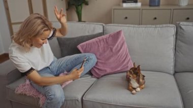 Sarışın genç bir kadın evcil köpeğini gri bir kanepede rahat bir oturma odasında cep telefonuyla fotoğraflıyor..