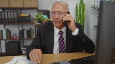 Takım elbiseli üst düzey bir beyaz adam ofisinde masasında çalışırken telefonla konuşuyor..