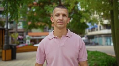 Pembe tişörtlü genç adam şehir parkında dikiliyor başparmağını kaldırıyor ve neşeli ifadelerle kaslarını gösteriyor.