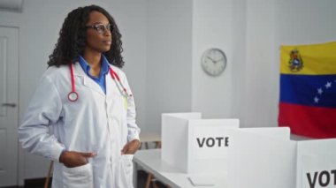 Venezuela seçim odasında, arka planda oy sandıkları, bayrağı ve saati olan kıvırcık saçlı kadın doktor.