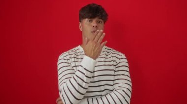 Genç İspanyol asıllı, çizgili tişört giyen, havada el ele öpüşen, güzel ve seksi bir ifade sergileyen, izole edilmiş kırmızı arka planda, kameraya sevgiyle bakan bir adam.