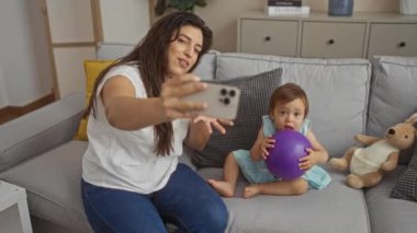 Anne, kızıyla birlikte evde oturma odasının iç dekorasyonunda mor topu tutarken selfie çekiyor.