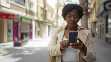 Orta yaşlı kıvırcık saçlı Afrikalı Amerikalı kadın güneşli bir sokak ortamında akıllı telefonuyla neşe içinde etkileşime geçiyor..