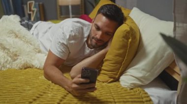 Sakallı, sarı bir yatakta uzanmış, modern bir yatak odasında akıllı telefon kullanan Latin kökenli genç bir adam..