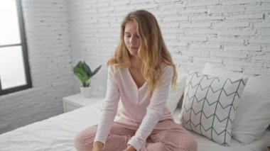 Genç bir kadın yatak odasının içinde, rahat bir yatakta oturuyor, bir cihaz tutuyor ve kulaklık takıyor.