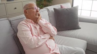 Gözlüklü ve pembe tişörtlü yaşlı beyaz bir adam, rahat bir oturma odasında, gri bir kanepede, yumuşak yastıklı ve kapalı bitkilerle dinleniyor..