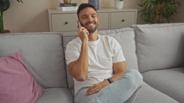 Genç, ispanyol, sakallı bir adam evde telefonla konuşuyor rahat bir şekilde bir kanepede oturuyor..