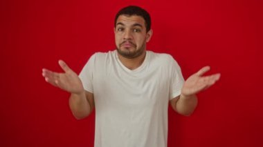 Beyaz tişört giyen genç İspanyol adam elleri havada ve kafası karışmış halde dikiliyor ve izole edilmiş kırmızı arka planda belirsizlik ifadesi var.