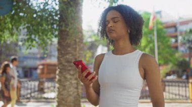 Kıvırcık saçlı güzel bir Afro-Amerikalı kadın güneşli bir günde şehir parkında akıllı telefon kullanıyor..