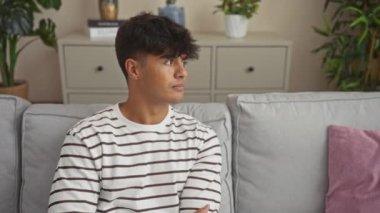 Çizgili gömlekli genç adam modern bir oturma odasında koltukta oturuyor, kameraya bakıp kollarını kavuşturuyor ve gülümsüyor..