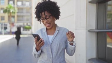 Genç, neşeli, kıvırcık saçlı ve gözlüklü Afro-Amerikalı bir kadın şehir caddesinde akıllı telefonuna bakarken kutlama yapıyor..