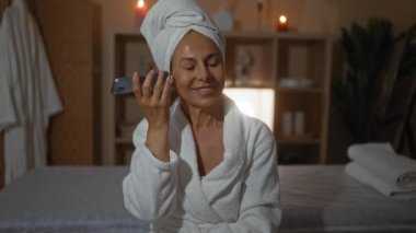 Kafasında bir havluyla spa odasında dinlenen, bornoz giymiş, telefon dinlerken gülümseyen, dingin bir sağlık merkezinde mum ışığıyla çevrili olgun bir kadın.