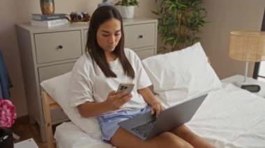 Yatak odasında dizüstü bilgisayar ve akıllı telefon kullanan bir kadın.