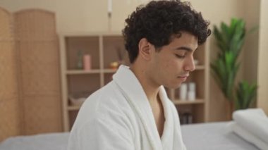 Genç bir İspanyol adam, arka planda bitki ve rafların olduğu kapalı bir ortamda beyaz bir bornoz giyerek bir kaplıca salonunda sağIık tedavisi görüyor..