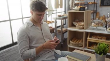 Fırında akıllı telefon kullanan ve arka planda ekmek ve pastalarla gülümseyen genç bir adam.