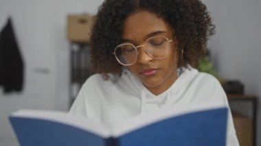 Kıvırcık saçlı ve gözlüklü genç Afrikalı Amerikalı kadın kapalı bir ofiste mavi bir kitap okuyor..