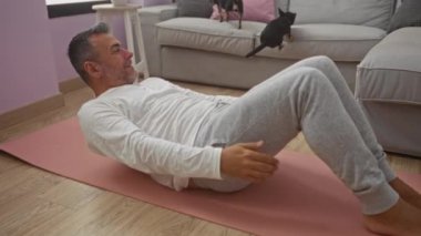 Orta yaşlı bir adam oturma odasındaki kanepede iki chihuahuayla yoga minderinde egzersiz yapıyor..