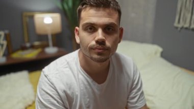 Sakallı, genç, yakışıklı bir İspanyol, rahat bir yatak odasında, arka planda güzel dekore edilmiş bir iç dekorasyonla kameraya samimi bir şekilde gülümsüyor..