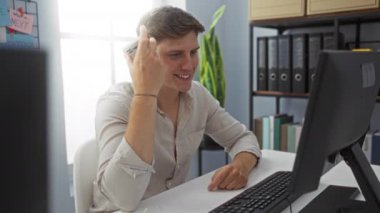 Akıllı telefondan konuşan ve ofiste bilgisayar monitörü ve arka planda klasörlerle çalışırken gülümseyen genç bir adam.