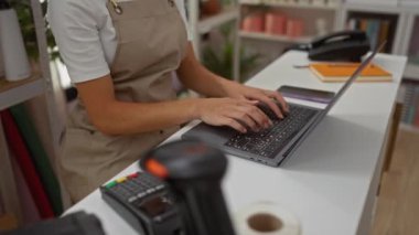 Ev dekorasyonu mağazasında dizüstü bilgisayarda çalışan kadının arka planda çeşitli ürünlerle dolu rafları var.