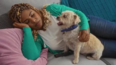 Saçları örgülü genç bir Afro-Amerikalı kadın rahat bir oturma odasında köpeğiyle kanepede dinleniyor..