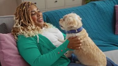 Saçları örgülü genç bir Afro-Amerikan kadın sıcak bir oturma odasında köpeğiyle vakit geçiriyor..