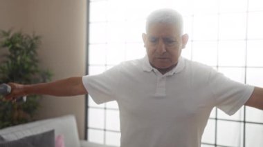 Orta yaşlı İspanyol bir adam oturma odasında dambıllarıyla egzersiz yapıyor, beyaz bir gömlek giyiyor, iyi ışıklandırılmış, modern bir iç mekanda bitkilerle ve rahat mobilyalarla kararlılık gösteriyor..