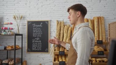 Bir fırında çalışan genç adam arka planda ekmek ve hamur işleriyle dolu raflarla...