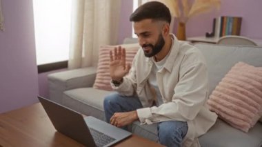 Sakallı genç İspanyol adam evdeki oturma odasından arıyor rahat dekorlu ve doğal ışıklandırmalı bir laptop kullanıyor.