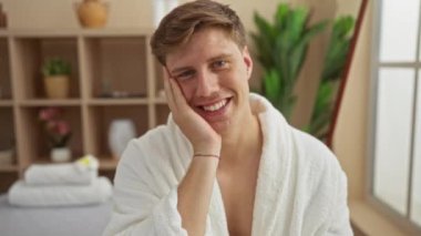 Kaplıcada beyaz bir bornoz giyen genç bir adam, bir sağlık merkezinde otururken gülümsüyor ve arka planda bir raf, kapalı alanda rahatlatıcı bir anın tadını çıkarıyor.