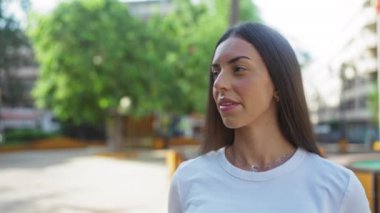 Güneşli bir günde arka planda ağaçlarla şehir parkında dikilen genç bir kadın çekici ve rahat bir tavır sergiliyor.