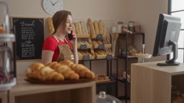 Ekmek ve hamur işleriyle çevrili bir fırında çalışırken telefonla konuşan genç bir kadın.