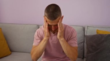 Genç adam oturma odasında bir kanepede oturuyor, pembe bir gömlek giyiyor ve yüzünü açık mor duvarlar ve renkli yastıklarla örten elleri ile stresli görünüyor.