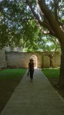 Palma de Mallorca 'da taş duvarlar ve ağaçlarla gölgeli bir yolda yürüyen bir kadın. İspanya şapkası takıyor ve bir çanta tutuyor.