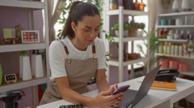 Dekoratörde telefon kullanan bir kadın, dizüstü bilgisayarının yanında önlük giyerken güzel düzenlenmiş ev dekorasyonu dükkanında, etrafta bir sürü ürünle birlikte.