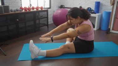 Genç bir İspanyol kadın spor salonundaki mavi bir paspasın üzerinde egzersiz yapıyor. Etrafı dambıl ve egzersiz topu gibi malzemelerle çevrili. Dövmelerini gösteriyor ve rahat bir spor kıyafeti giyiyor..