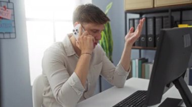 Akıllı telefondan konuşan ve ofis çalışma alanında bilgisayar ve kitaplıkla el kol hareketi yapan genç adam.