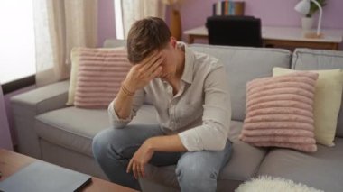 Oturma odasındaki bir kanepede oturan genç adam endişeli ve stresli görünüyor. Alnında yastıklar ve ahşap masadaki bir dizüstü bilgisayarla.