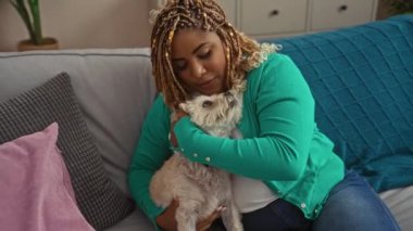 Saçları örgülü genç bir Afrikalı Amerikalı kadın sıcak bir oturma odasında köpeğine sarılıyor..