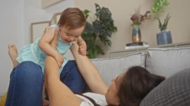Yeni yürümeye başlayan kızıyla modern bir salonda rahat bir kanepede oynayan bir kadın, neşeli aile bağlarını ve aşklarını dekor ve bitkilerle dolu rahat bir evde sergiliyor..