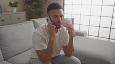 Sakallı genç bir İspanyol adam oturma odasındaki gri bir koltukta oturur, akıllı telefonuyla ciddi ciddi konuşur, etrafı kapalı bitkiler ve modern dekorlarla çevrilidir..