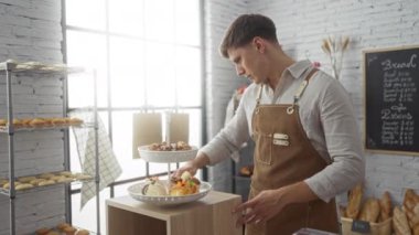 Fırında önlük giyen genç bir adam arka planda çeşitli ekmek ve menülerle kapalı bir dükkânda hamur işleri düzenliyor.