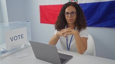 Dominik Cumhuriyeti bayrağı önünde dizüstü bilgisayarı ve oy sandığıyla masada oturan bir kadın.