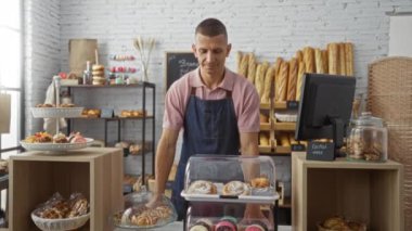 Bir fırında çalışan genç adam önlük ve pembe polo tişört giyiyor. Etrafı çeşitli ekmek ve hamur işleriyle çevrili..