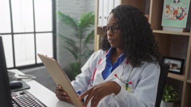 Genç, kıvırcık saçlı, beyaz önlüklü, gözlüklü ve klinikte oturan Afro-Amerikalı bir kadın doktor, iş yerinde dikkatlice bir pano okuyor..