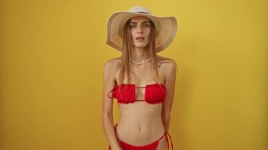 Bikinili ve yaz şapkalı kadın izole edilmiş sarı arka planda sessizce el kol geziyor, beyaz kadın dudaklarında parmaklarıyla sessizlik işareti yapıyor, yaz kıyafetinde sır saklıyor.