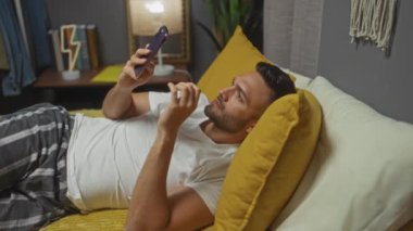 Latin kökenli genç bir adam rahat bir yatak odasında, bir yatakta uzanmış akıllı telefonuna bakıyor..
