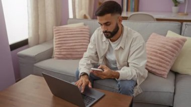 Genç adam, pembe yastıklı, bej perdeli ve ahşap masalı rahat bir oturma odasında kanepede otururken akıllı telefon ve dizüstü bilgisayar kullanıyor.