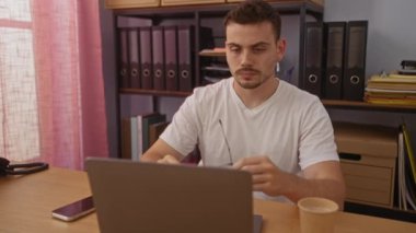 Sakallı ve gözlüklü genç bir İspanyol iş yerinde oturuyor, etrafı dosya raflarıyla çevrili ve bilgisayarında akıllı telefon ve kahve bardağıyla çalışıyor..