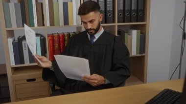 Yargıç cüppeli genç İspanyol adam ofis odasında kitap rafı bilgisayarı ve arka planda hukuk kitaplarıyla doküman okuyor.